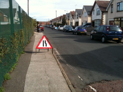 A 'road narrows' sign obstructs the pavement